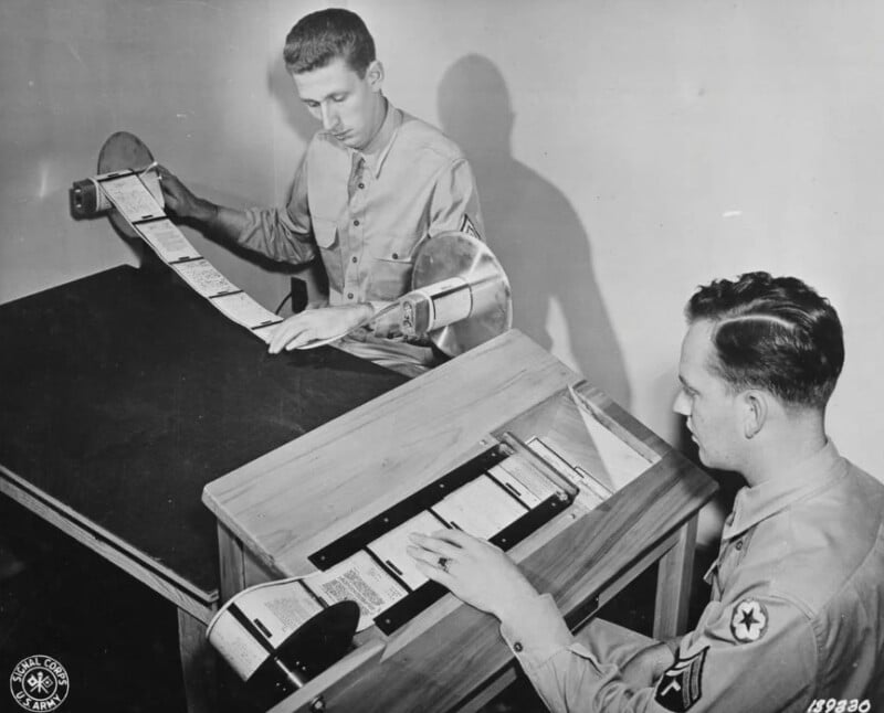 Two men in military uniforms are operating early computing equipment. One is feeding paper tape into a machine, while the other adjusts it. They are seated at a wooden desk with a reel of tape. The image is in black and white.